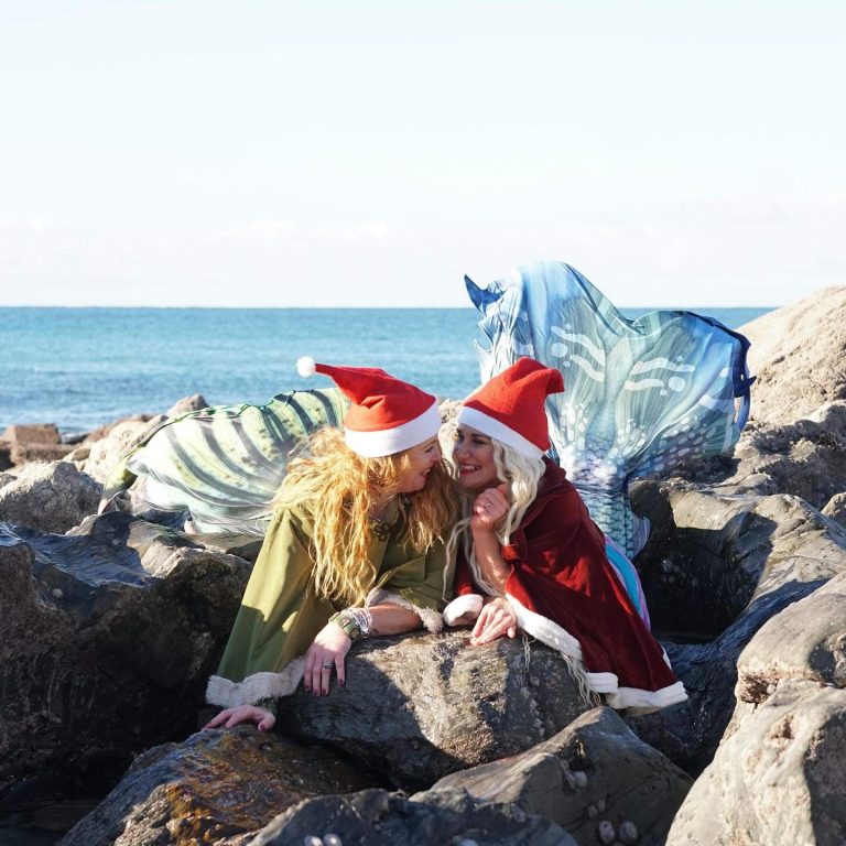 2 women dressed as mermaids lying on a beach wearing xmas hate.