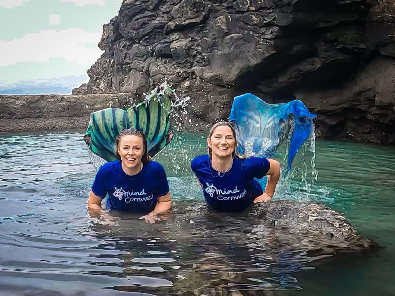 Two women with mermaid tails, in the sea smiling and wearing Cornwall Mind t shirts.